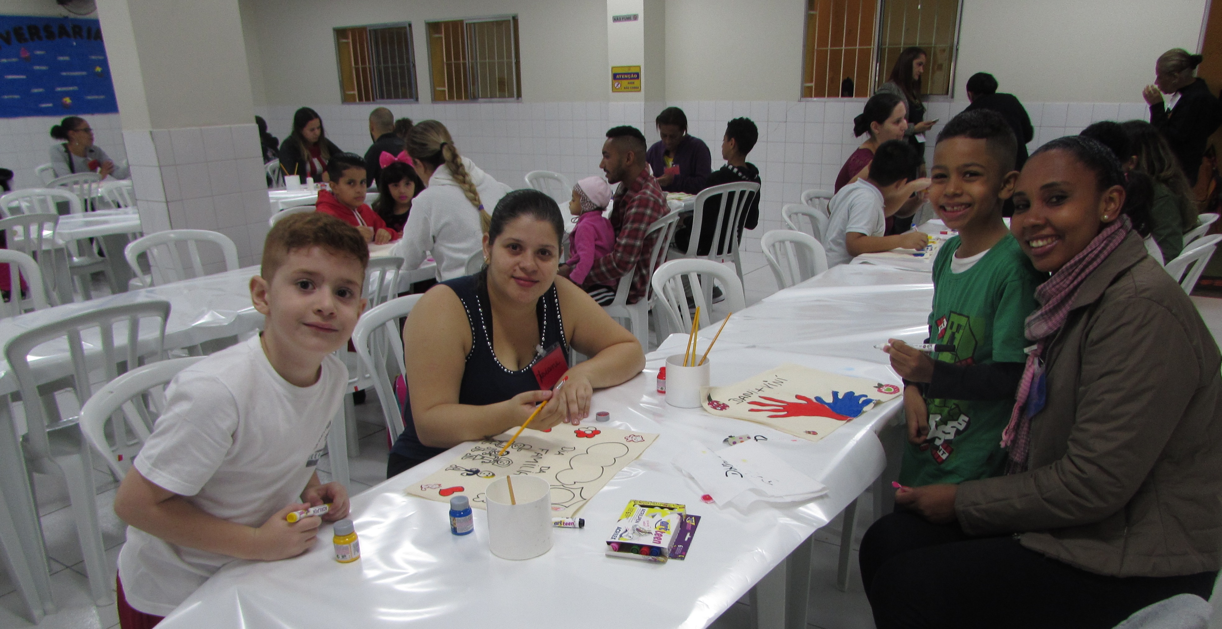 Em um espaço fechado, duas mães posam com seus filhos meninos enquanto estão fazendo atividades artísticas em uma mesa branca. Ao fundo estão sentadas as outras famílias