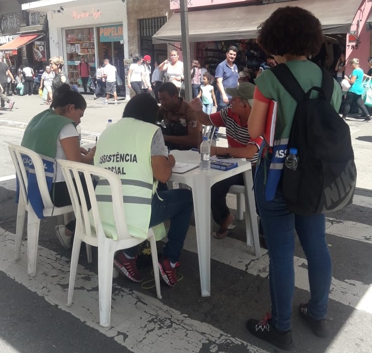 Com uma tenda montada na rua, orientadores socioeducativos e assistentes sociais com coletes da Assistência Social (cores azul, verde e verde água) atendem dois homens em uma mesa. 