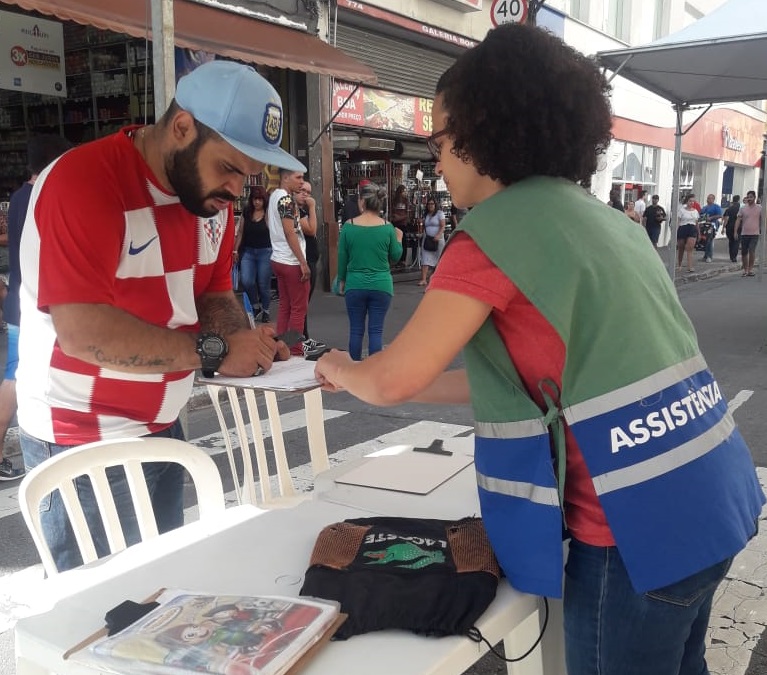 Um homem com boné azul e camiseta de esporte vermelha e branca, preenche um papel em uma prancheta com a ajuda da assistente social.