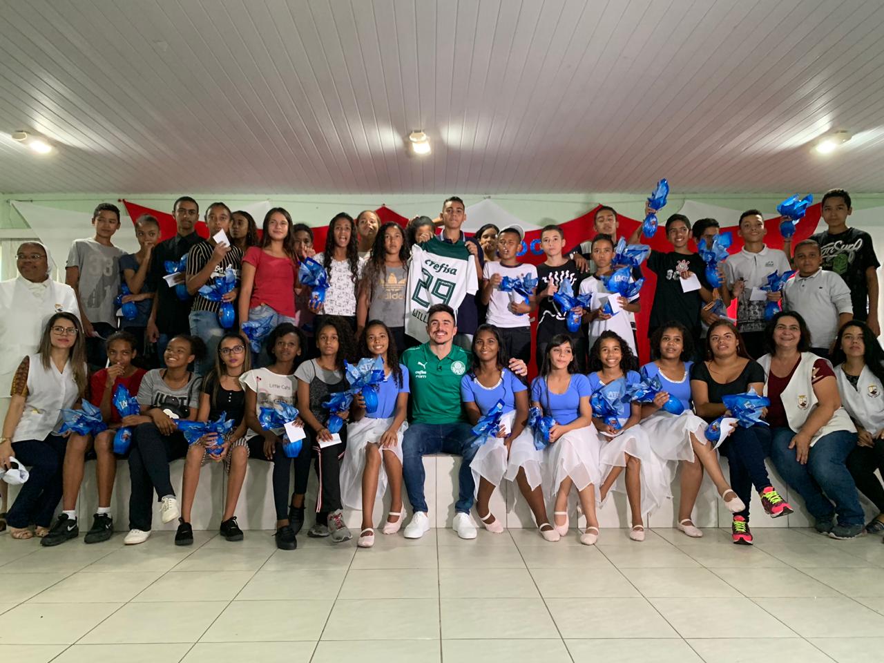 Dentro de uma sala e à frente de um palco, crianças e adolescentes do CCA IASE posam com o ovo de páscoa para foto com o jogador do Palmeiras Willian. Na imagem também estão funcionários do serviço.