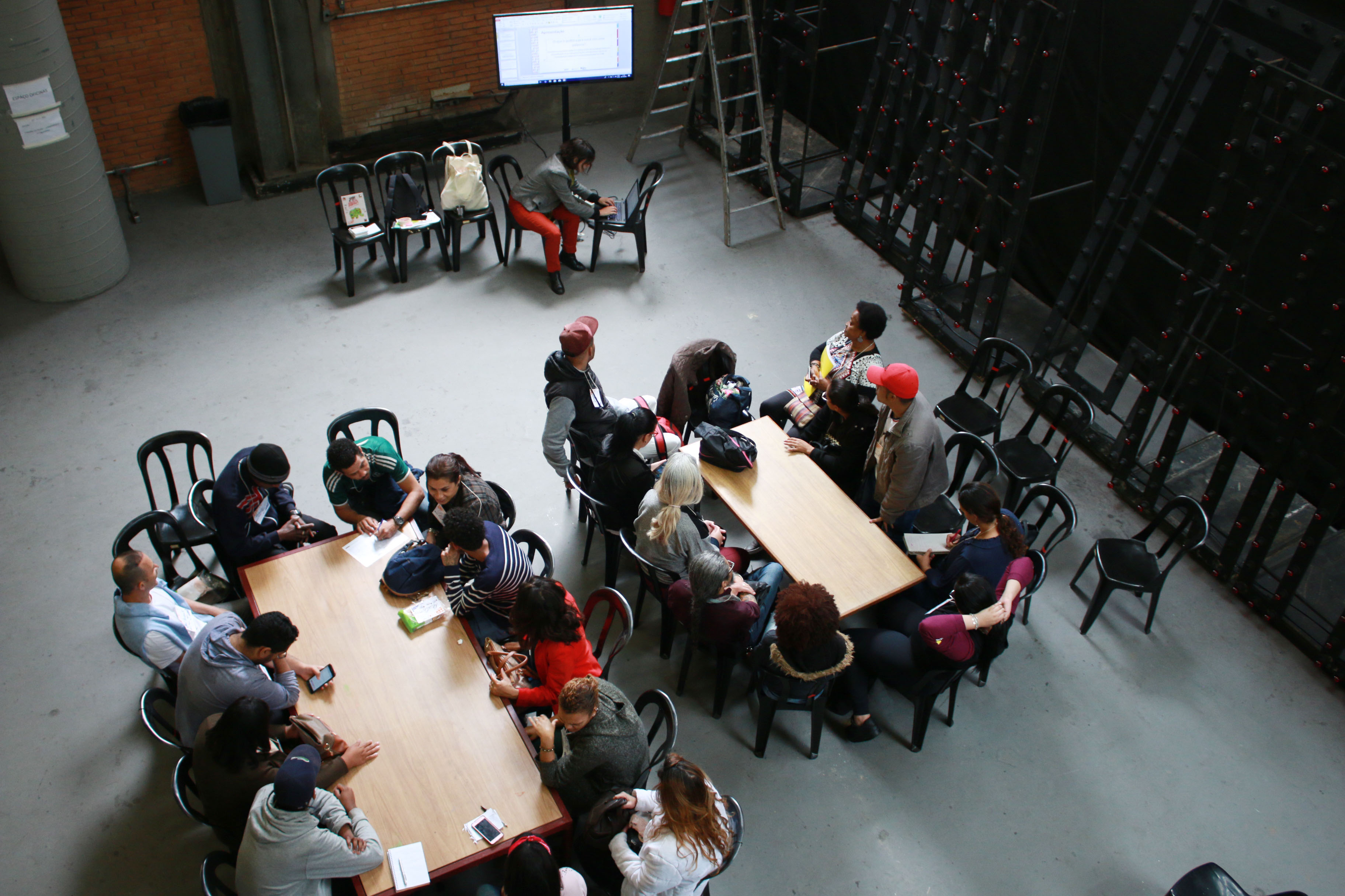 Vinte e duas pessoas divididas em duas mesas de maneiras realizam atividade com papéis e calculadora. Separada do grupo, uma mulher mexe em notebook conectado a uma tela.