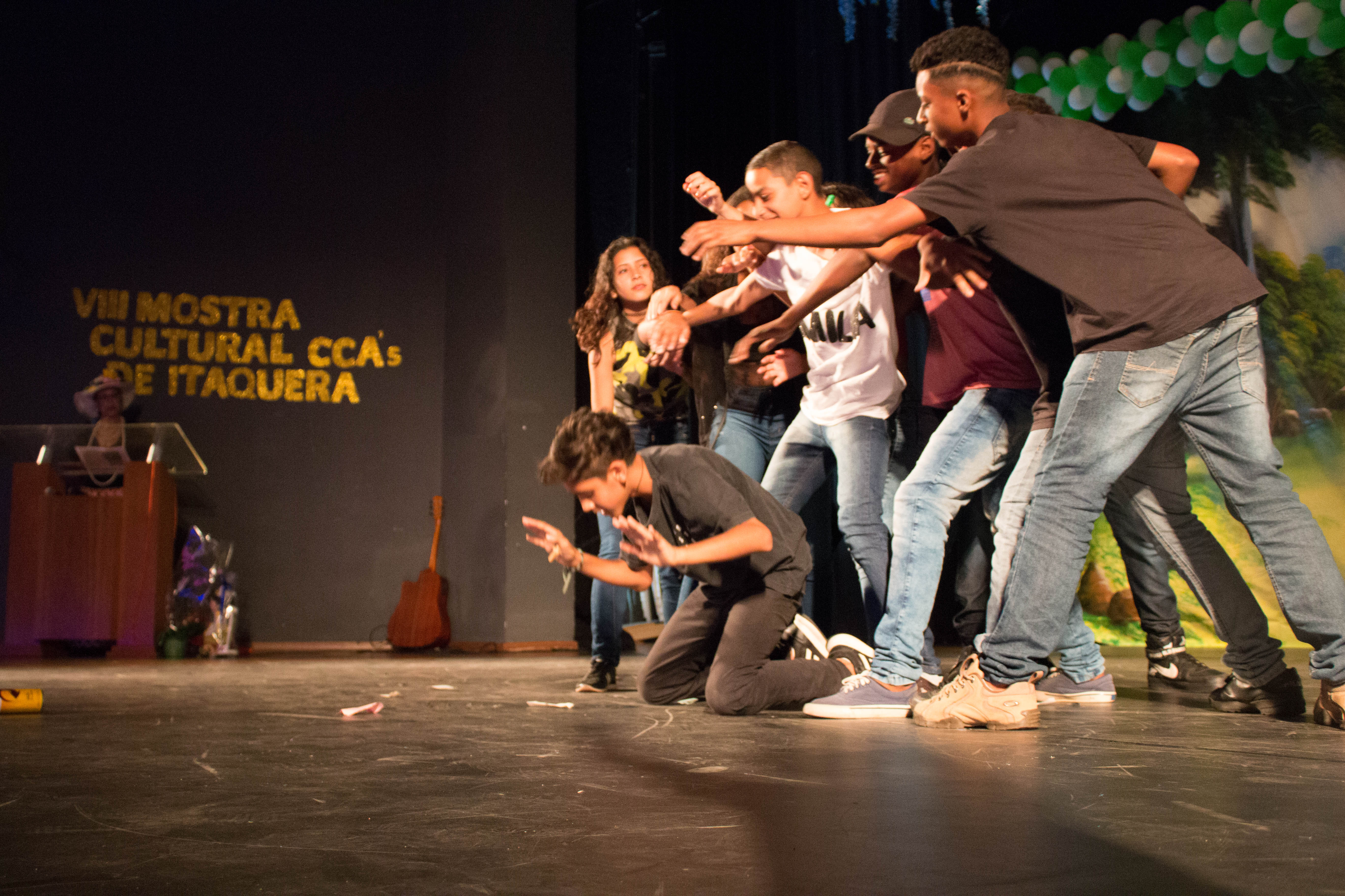 Sete crianças e adolescente fotografados de lado durante apresentação de dança em um palco preto. Ao fundo há uma mulher em um palanque. Atrás dela, em letras amarelas “VIII Mostra Cultural CCAs de Itaquera”.