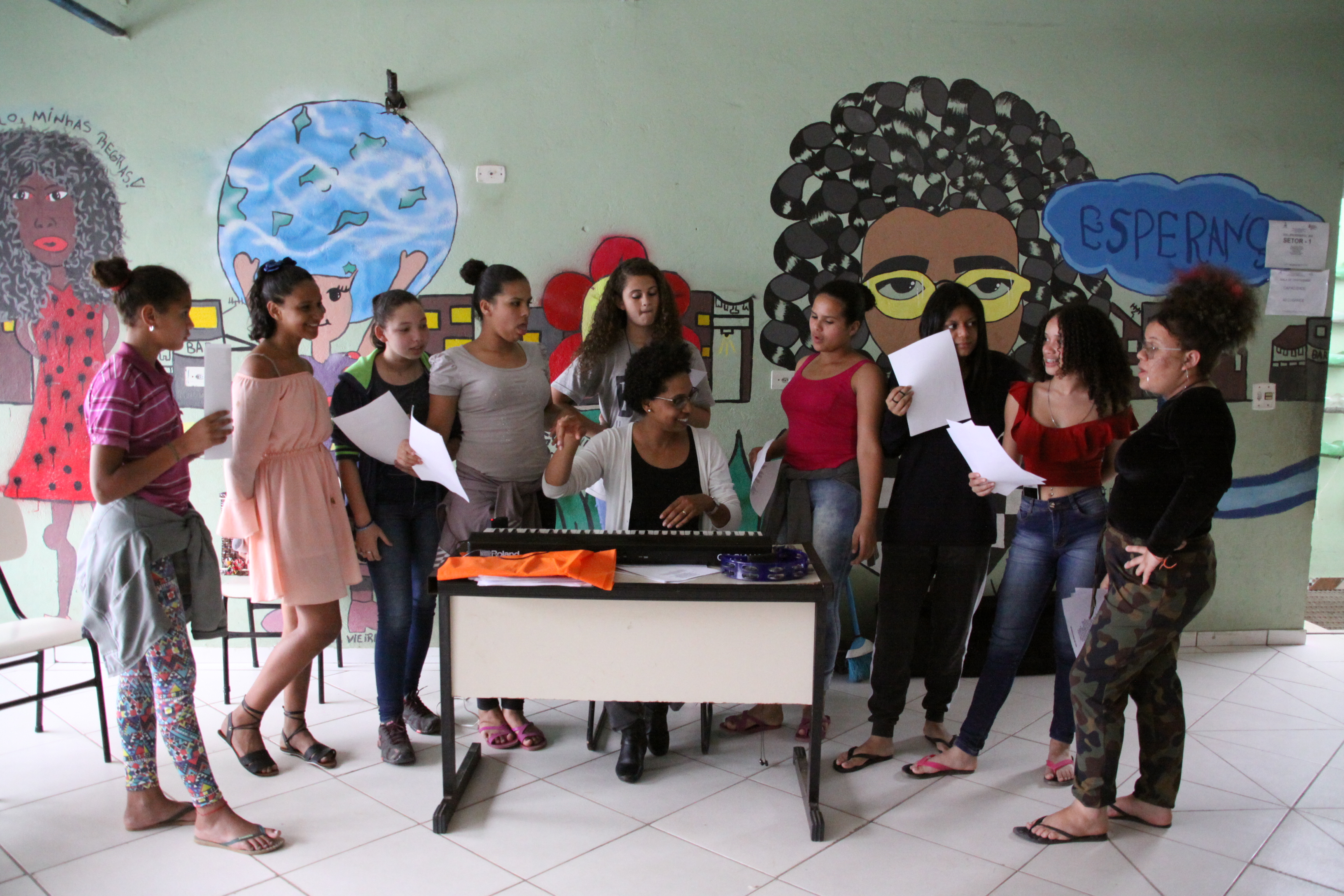 Nove garotas estão de pé dentro de uma sala, uma do lado da outra. Todas observam a professora que está sentada, tocando um teclado em cima da mesa. Algumas garotas seguram papéis em suas mãos