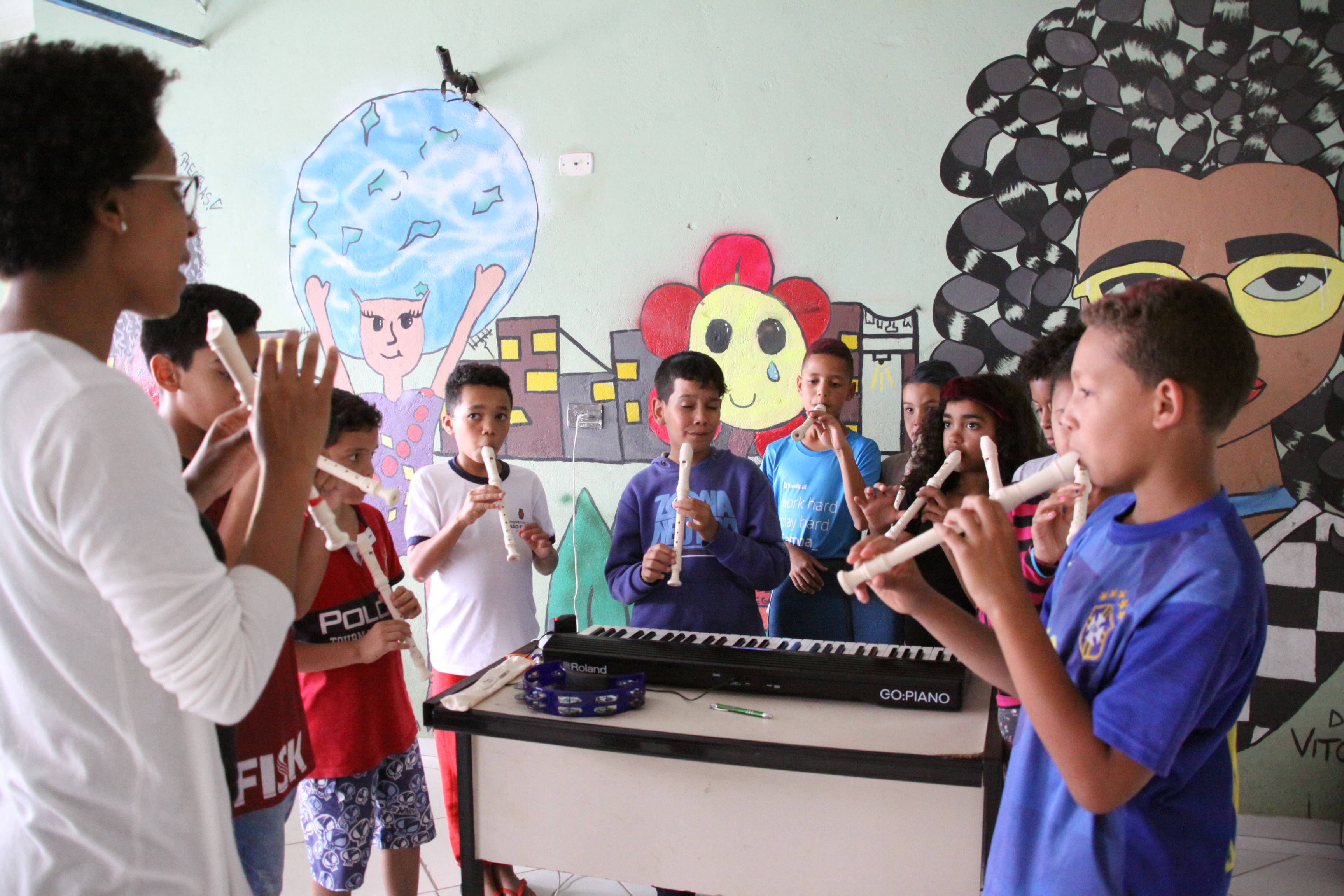 Dez crianças, estão de pé, dentro de uma sala, lado a lado. Todos estão com flautas na mão e estão tocando o instrumento. Ao lado deles, há uma professora, com uma flauta na mão, dando instruções para os alunos.