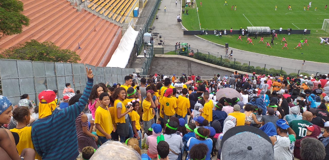 Da arquibancada do estádio, crianças do CCA assistem à partida. Algumas estão de costas para a foto e outras olham diretamente. 