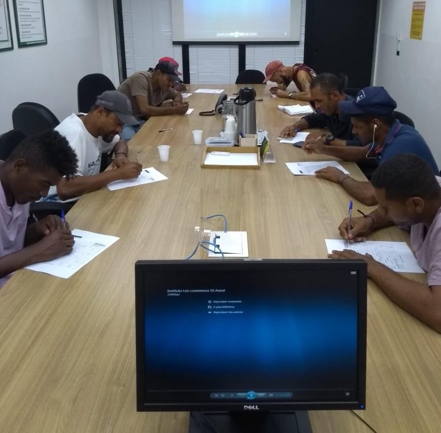 Oito homens estão dentro de uma sala, sentados em cadeiras, escrevendo com canetas em um papel branco que está sobre uma mesa de madeira. No centro da mesa há uma jarra de café, de água, copos e açúcar.