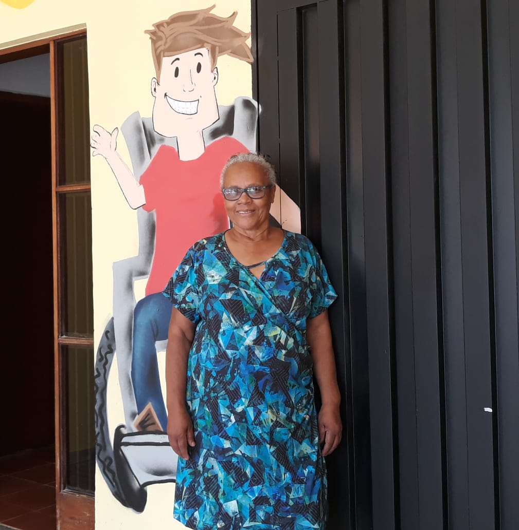 Dona Nadir, senhora negra com cabelos brancos e óculos de grau, veste um vestido azul enquanto posa para foto. Ela está na frente de uma parede com  desenho de um menino cadeirante.