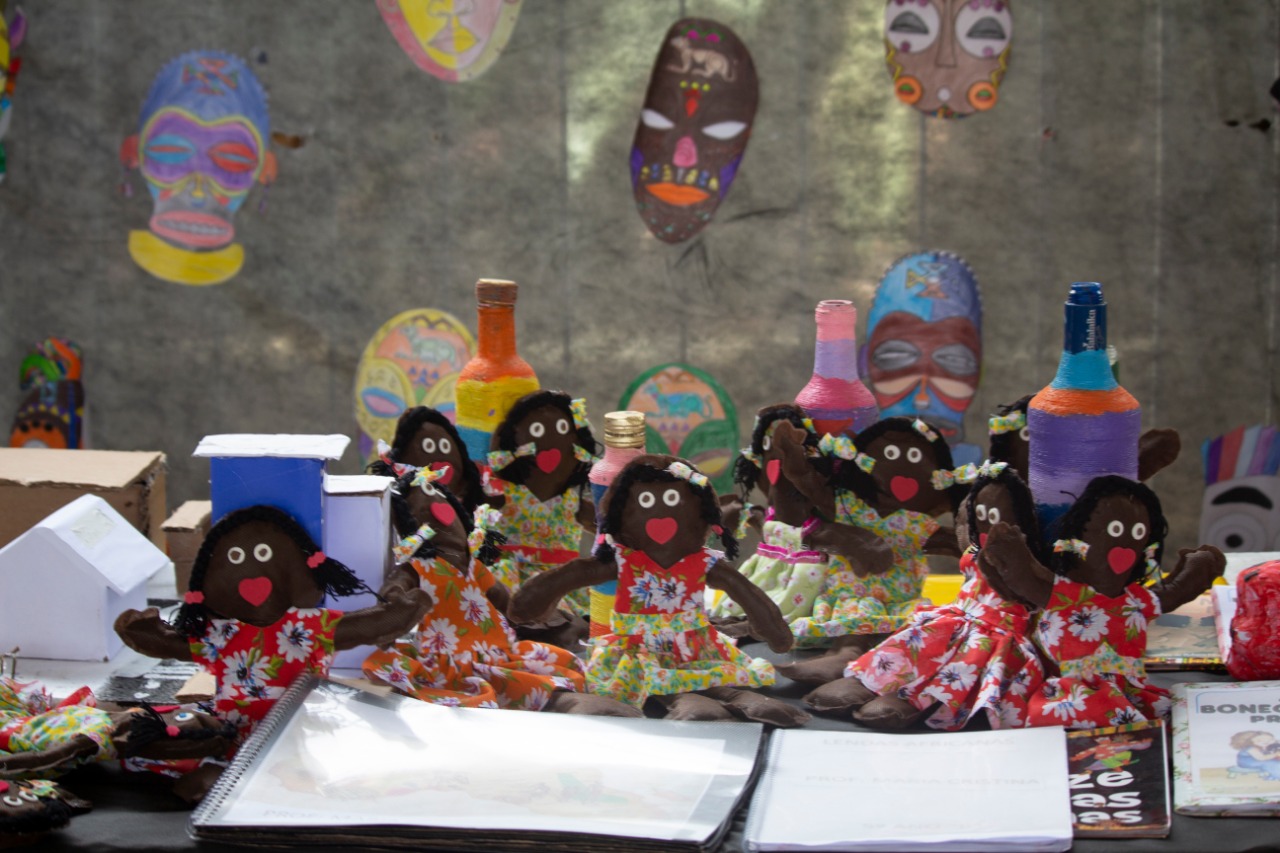 Bonecas negras de pano com vestidos coloridos e floridos, feitos por conviventes dos serviços, apoiadas em uma mesa em exposição.