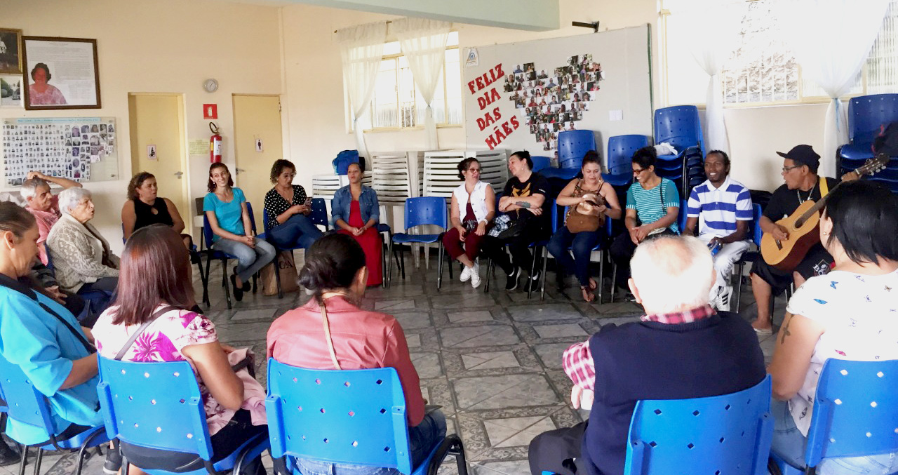 Em círculo, pessoas de várias idades interagem  em uma roda de conversa e alguns homens tocam violão e pandeiro