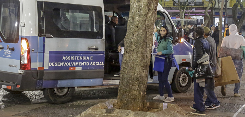 Durante a noite no centro da cidade, uma van da assistência social está parada enquanto a orientadora socioeducativa com colete verde e azul atende homens em situação de rua