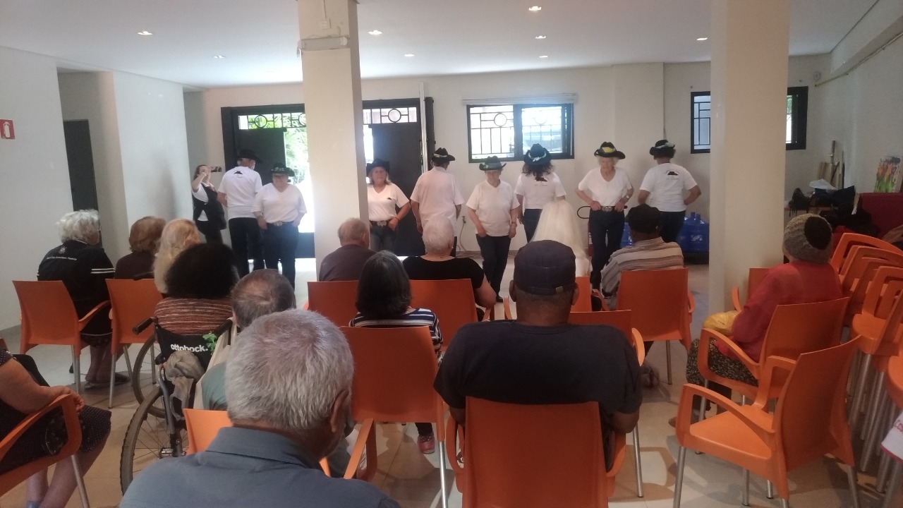 Dezenas de idosos, que estão sentados em cadeiras laranjas, observam oito idosos que estão fazendo uma coreografia.
