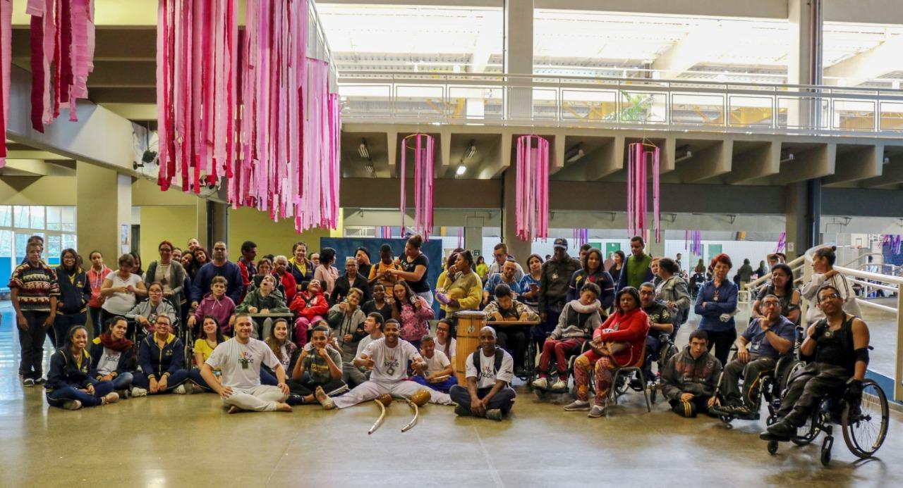 Centenas de pessoas estão posando para a foto, alguns estão de pé, alguns sentados, e outros na cadeira de rodas. Há fitas rosas no teto enfeitando a parte de cima do local.