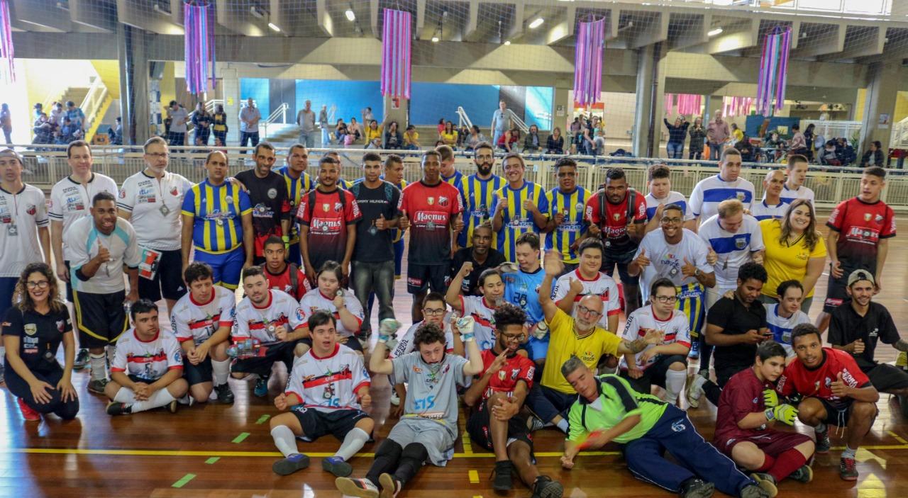 Quarenta e seis pessoas posam para a foto, no meio da quadra de futsal. Jogadores de ambos os time, comissões técnicas e os árbitros. Alguns estão de pé e outros sentados. Na parte de trás, há uma arquibancada com algumas pessoas.