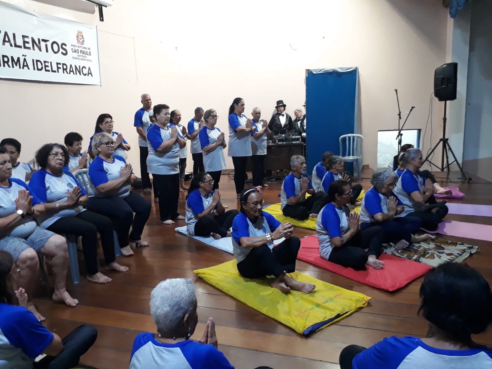 Dezenas de idosos estão em cima de um palco, usando camisetas brancas com mangas azuis. Todos estão sentados, com as palmas das mãos juntas, numa prática de yoga.