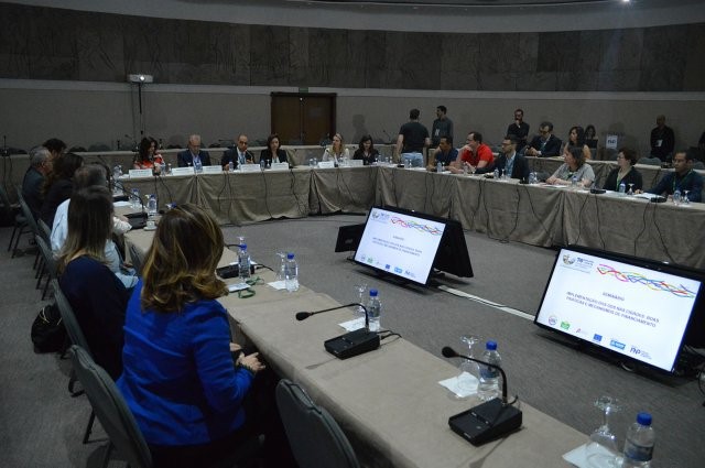 Participantes do evento, sentados em mesas, formam uma roda. No meio estão algumas telas de televisão exibindo conteúdo da reunião.