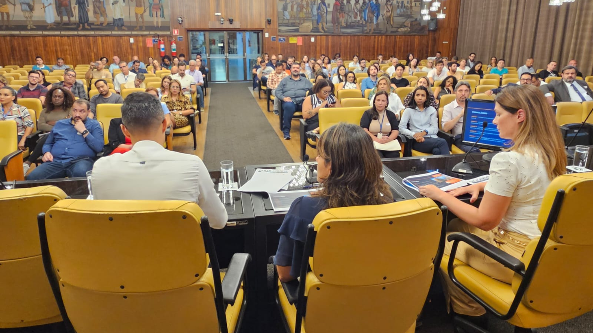 Fotografia apresenta de frente um auditório com pessoas prestando atenção na apresentação.