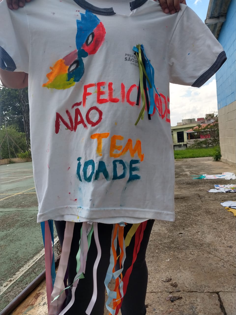 : A imagem mostra uma camiseta branca personalizada para o carnaval. No centro está escrita a frase “Felicidade não tem idade”.