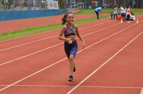 Na imagem, criança correndo sorridente na pista de atletismo.
