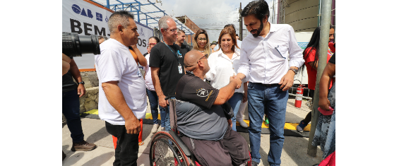 Prefeito e secretários no Mutirão - Inclui Sampa, na cidade Tiradentes. Prefeito segura a mão de um rapaz que está em uma cadeira de rodas.