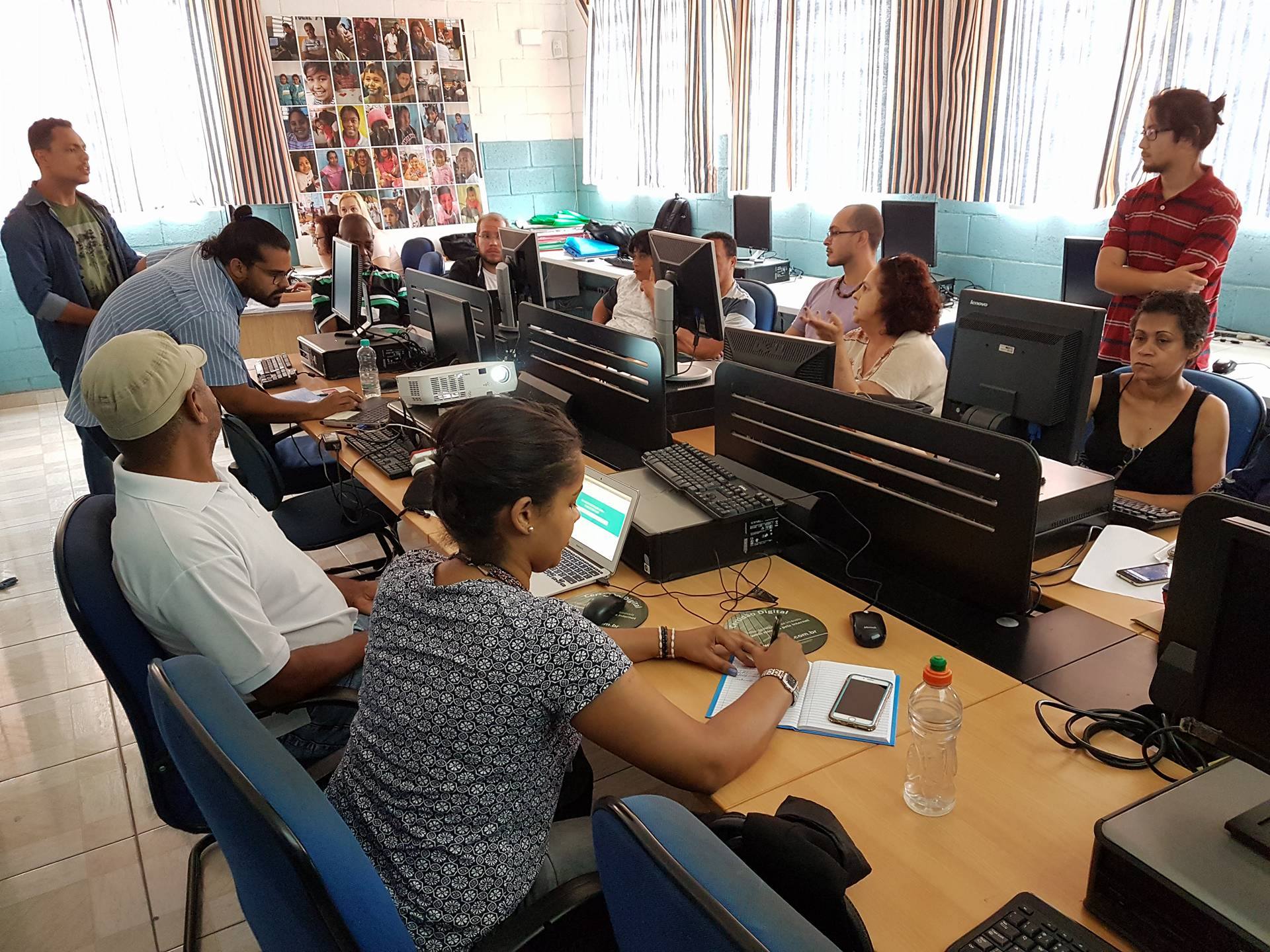 Laboratório de computação, membros do conselho gestor sentados nos computadores e instrutores da CGM em pé explicando os temas.