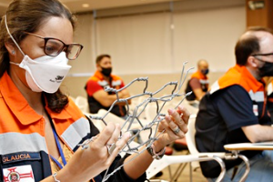 #PRATODOSVEREM- em uma sala uma mulher com óculos e mascara branca, uniforme da defesa civil, nas cores, azul,branco e laranja, segura uma armação de arame.  Ao fundo três homens com o mesmo uniforme e máscara , olham para frente.