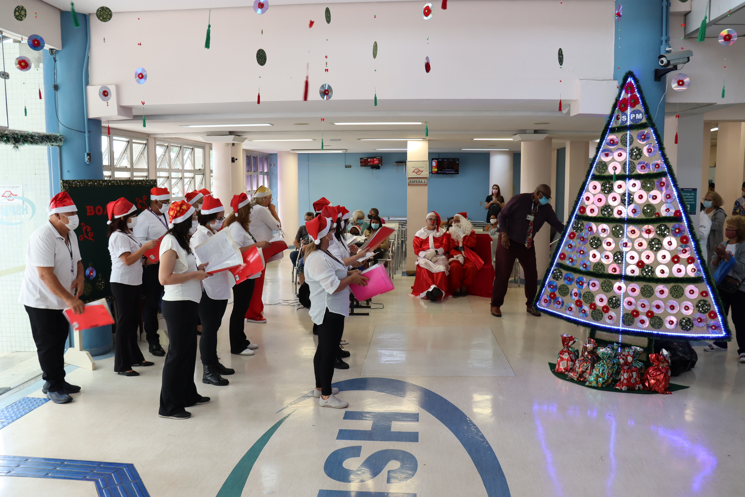 Foto panorâmica do saguão principal do HSPM com grupo de pessoas do coral, uniformizadas e usando gorro do Papai Noel, de pé e cantando.