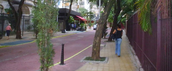 Foto de mulher, de costas, andando em calçada de cor amarela. Há uma árvore na calçada e, ao lado, rua pintada na cor vermelha.