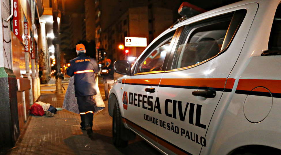 Foto mostra agente da Defesa Civil nas ruas da cidade entregando cobertor aos moradores de rua.