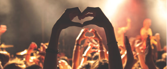 #PraCegoVer Foto de plateia em show musical mostra várias braços e mãos em sequência em forma de corações. A foto está em tons amarelos