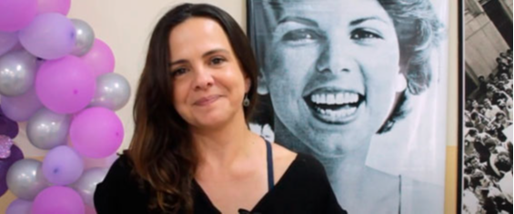 #PraTodosVerem Lili Grammont, filha de Eliane Grammont, comemora o Centro de Referência da Mulher que tem o nome de sua mãe. É uma mulher de cabelos pretos e na foto está à frente de um mural como a foto de Eliane