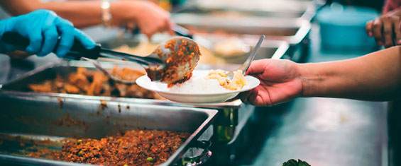 Uma mão segurando uma colher e servindo carne moída em um prato com arroz e que está sendo segurado pela mão de outra pessoa.