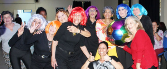 Foto de mulheres em pé, vestidas de preto e usando perucas coloridas. uma delas segura uma bola grande colorida.