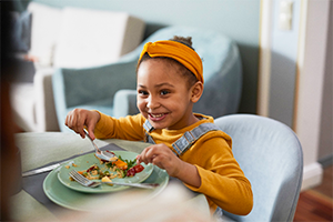 Criança negra, vestida com com blusa de manga comprida mostarda, tiara da mesma cor e macacão. Ela sorri e come comida de uma prato verde, posto sobre uma mesa a sua frente.