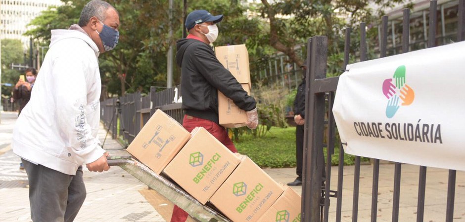 Foto de dois homens usando máscaras e entregando cestas básicas para a população em situação vulnerável.