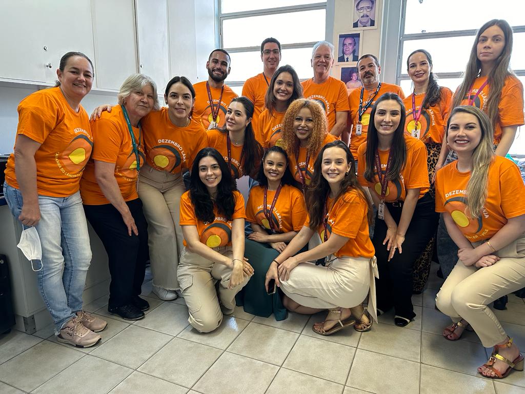 Fotografia na clínica de dermatologia do HSPM com a equipe composta por 17 pessoas, que estão vestidas com camisetas laranjas com o logo da campanha estampado.