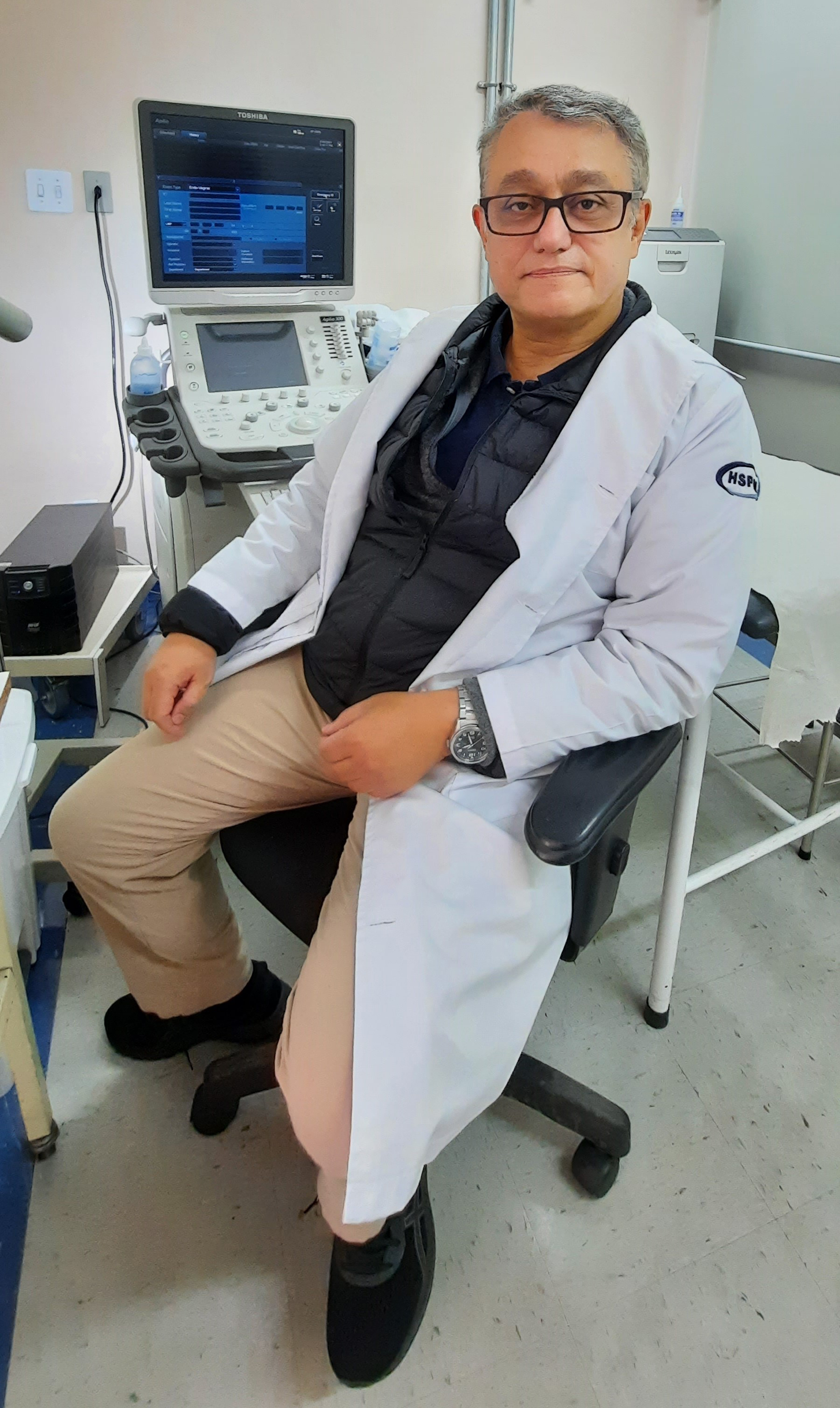 Foto do médico, de jaleco branco e óculos, sentado em frente a uma equipamento de ultrassom