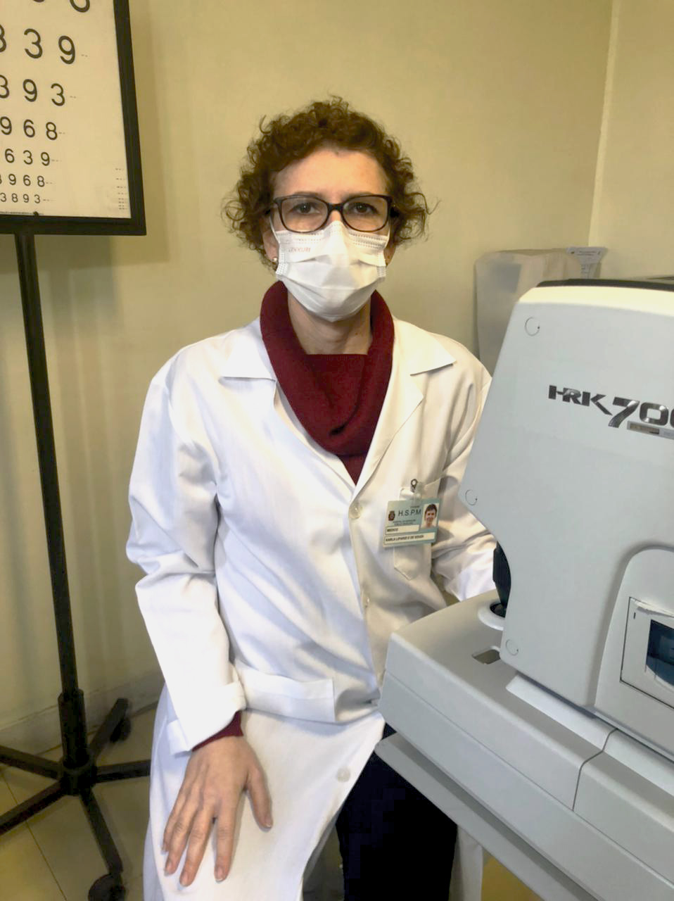 Foto da médica, com avental branca, máscara e óculos, sentada em frente a um aparelho oftalmológico