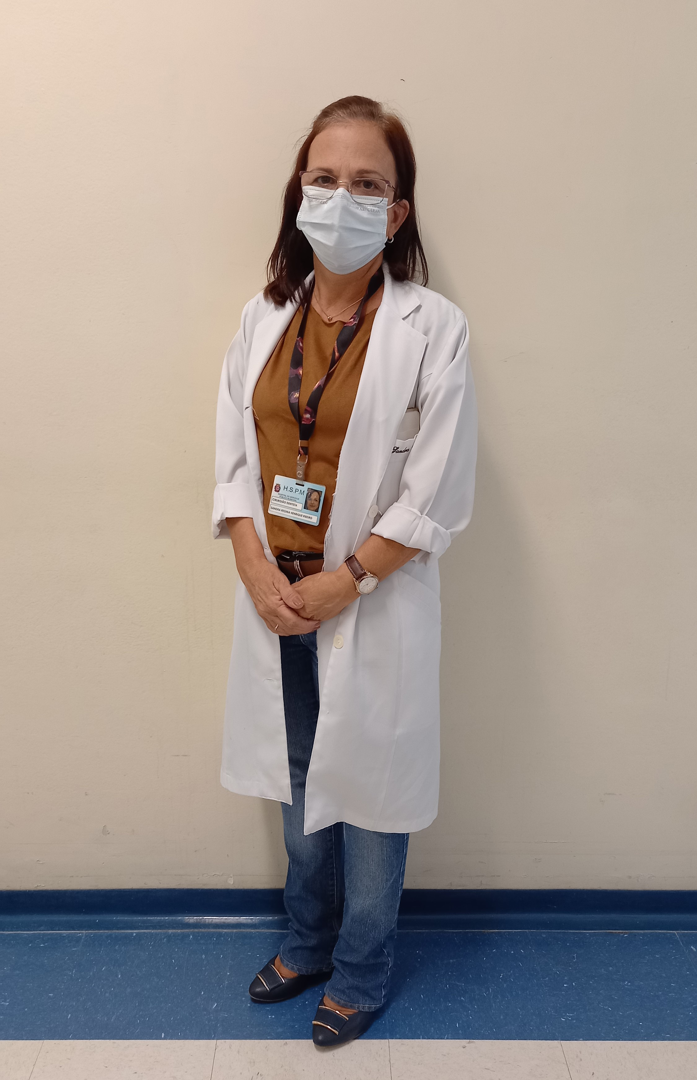 Foto da Dra. Sandra em pé em frente a uma parede bege e piso azul