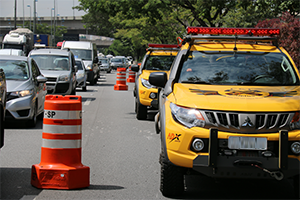 Fotografia com duas viaturas da CET à direita, cones laranja ao meio e carros transitando na via ao lado esquerdo