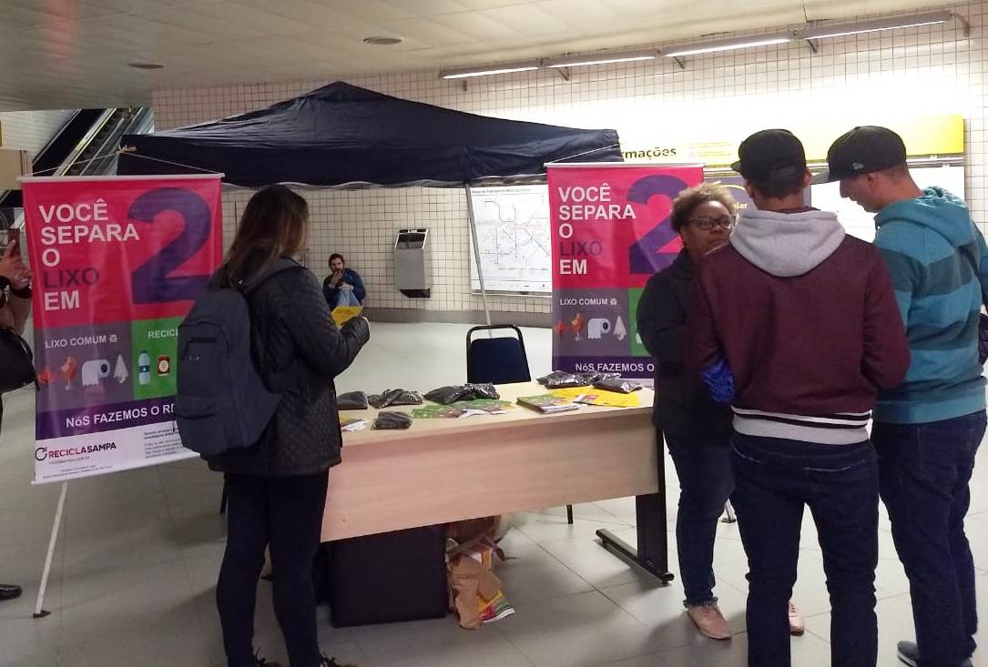 Pessoas reunidas na tenda de divulgação do Recicla Sampa na linha 4-Amarela. Em cima da mesa observamos sacos de composto.