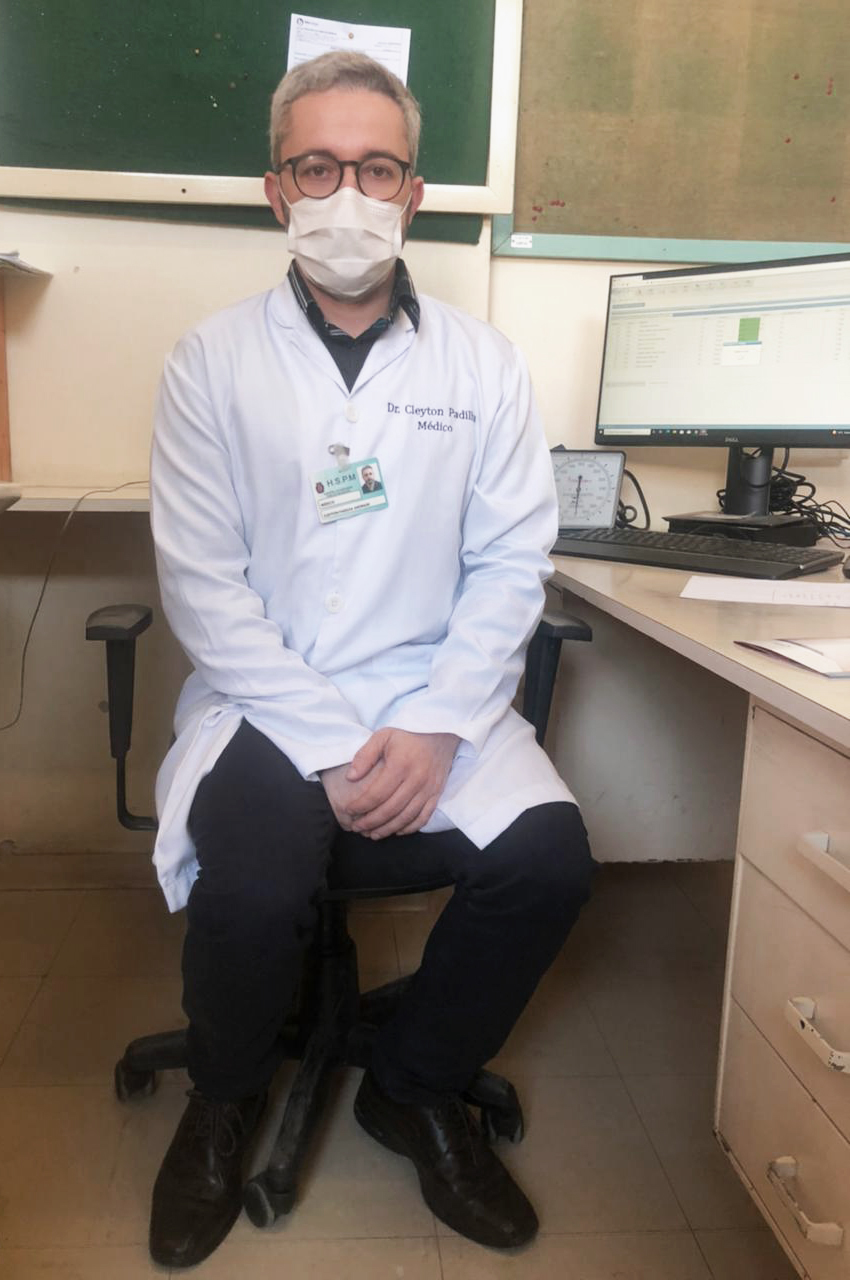 Foto do médico, com jaleco branco, sentado em frente a um computador