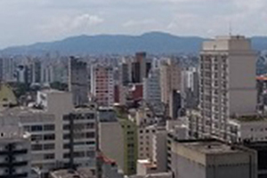 Vista panorâmica da cidade de São Paulo, sob um céu azul com algumas nuvens há diversos prédios, e ao fundo algumas colinas.