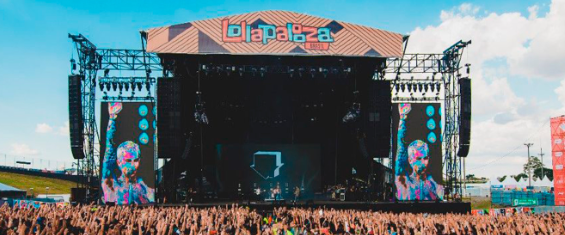 Foto apresenta um palco colorido, com Lollapalooza escrito acima e a plateia com as mãos levantadas embaixo