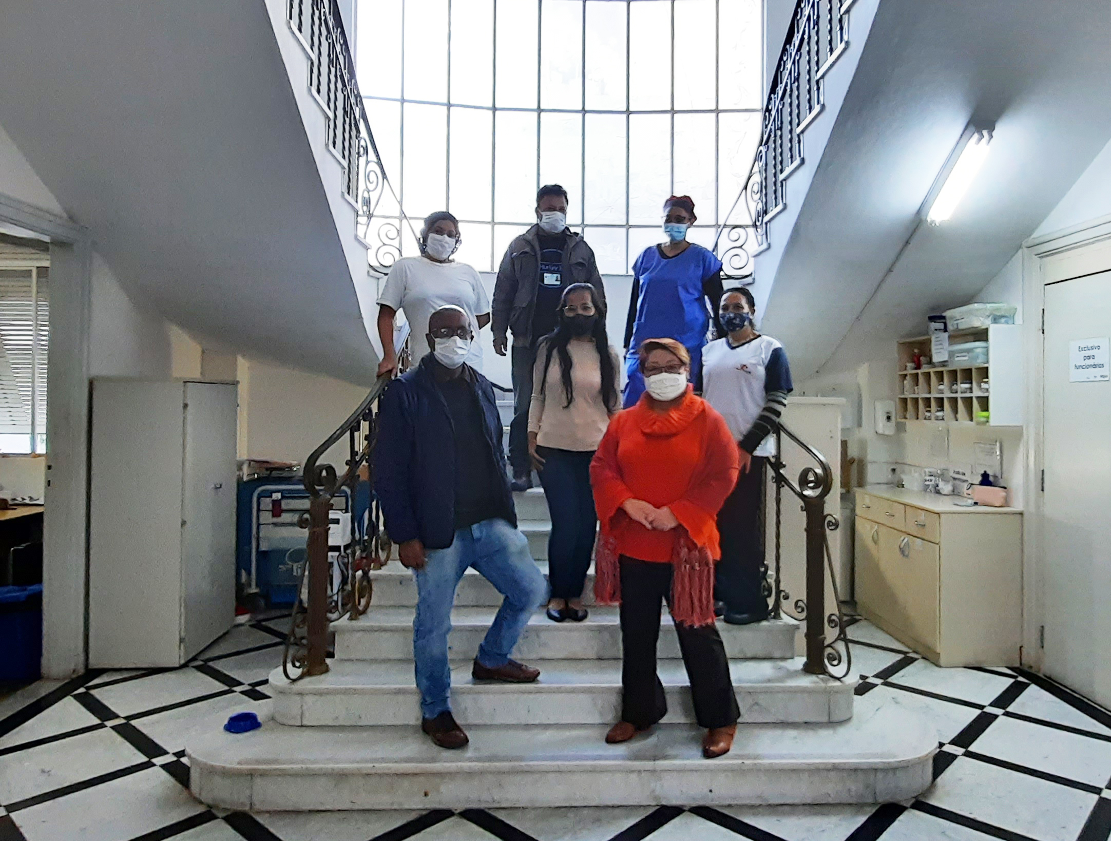 Foto de parte da equipe da hospedaria, sete pessoas em pé em uma escada que tem uma janela grande ao fundo