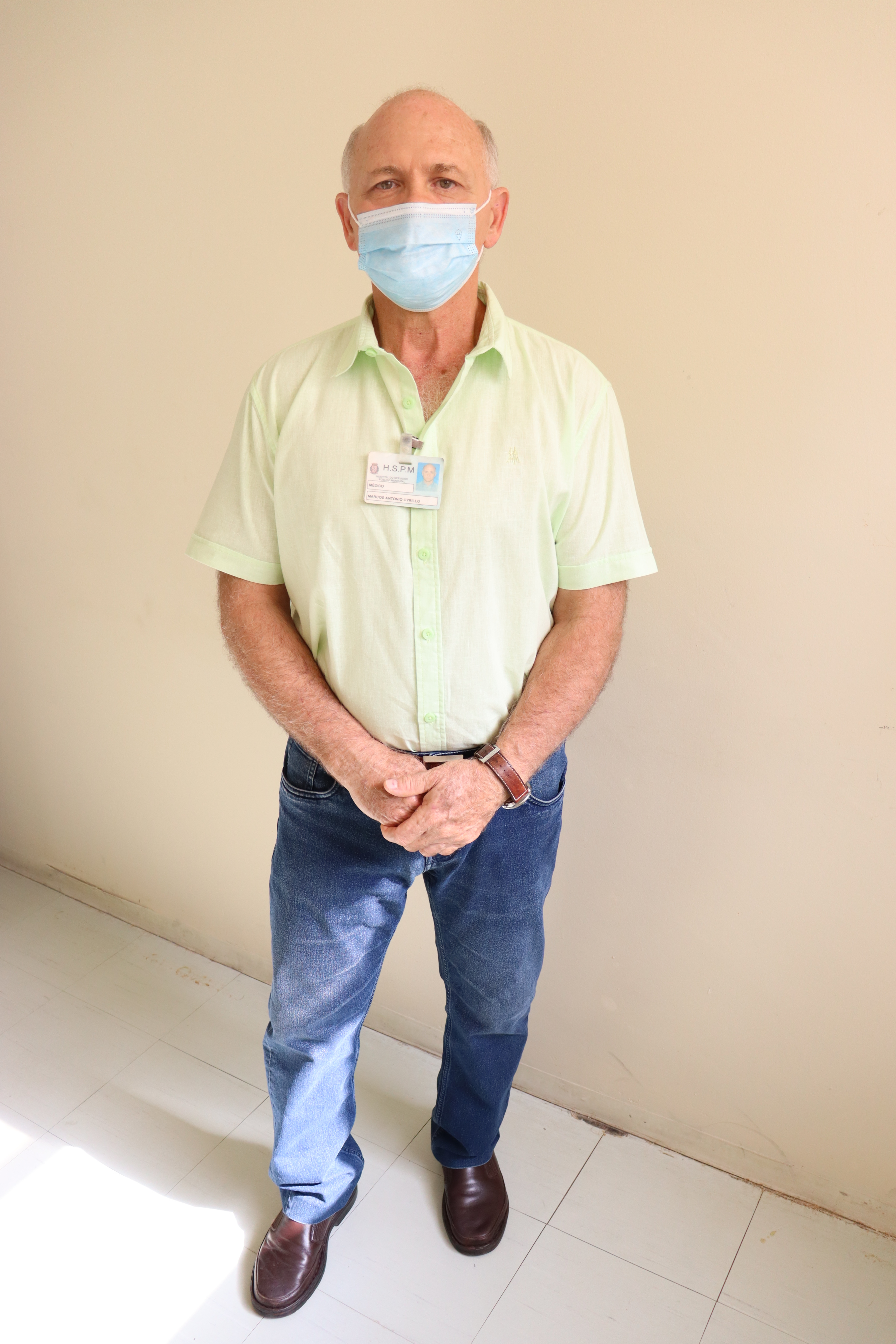 Foto do dr. Cyrillo, de sapatos escuros, calça jeans, camisa verde e máscara