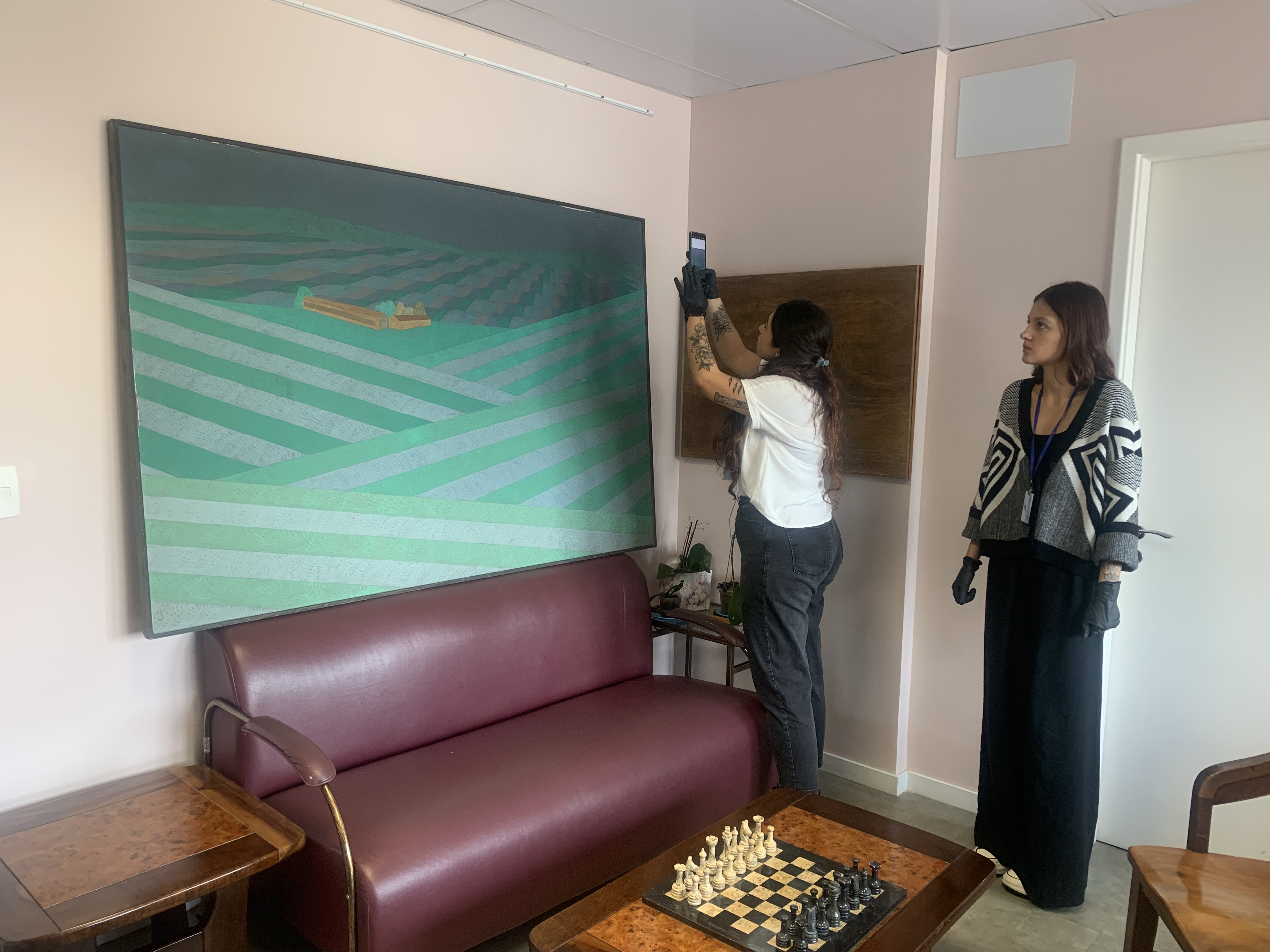 Fotografia na sala da Superintendência do HSPM. Aparecem duas profissionais conservadoras olhando e analisando uma das cinco obras de arte. Uma delas utiliza um celular para fotografar o quadro.