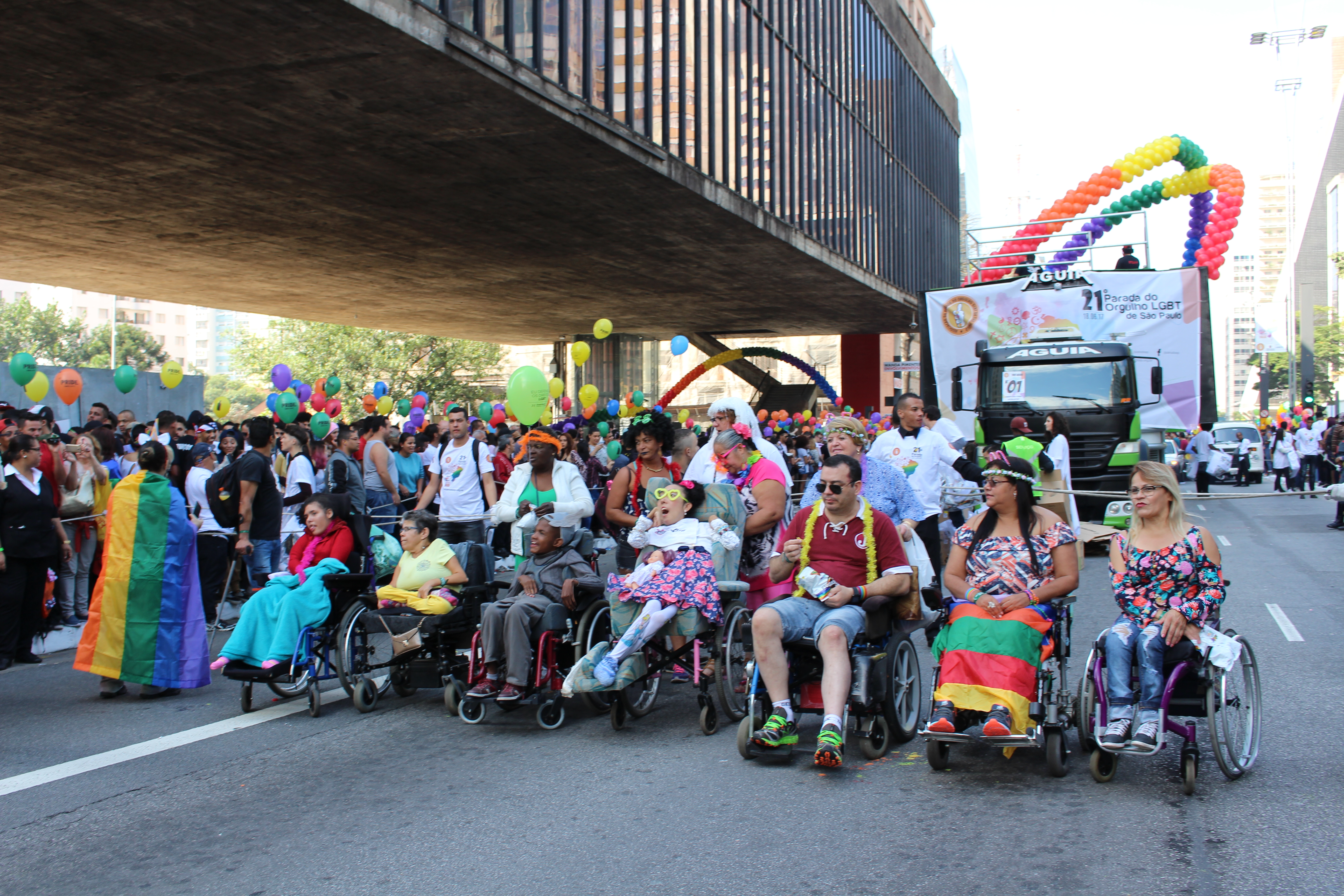 Pessoas em cadeira de rodas, com mobilidade reduzida e demais deficiências reunidas na Avenida Paulista na Parada do Orgulho LGBT. Há diversos balões coloridos.