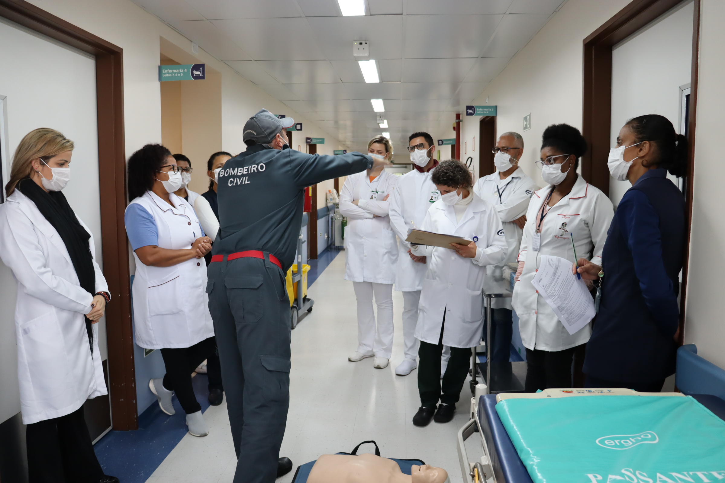 Imagem panorâmica da equipe no corredor do hospital em pé recebendo as instruções do bombeiro civil, que gesticula apontando uma direção