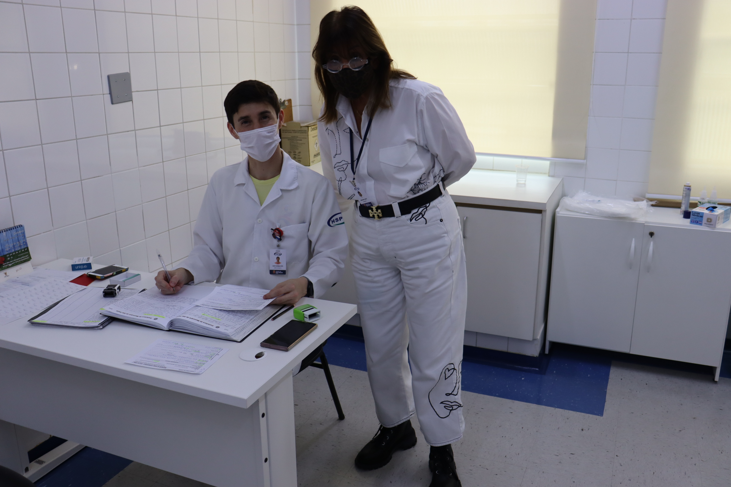 Foto de uma sala de atendimento, com instrutora em pé e enfermeiro sentado à mesa com documentos