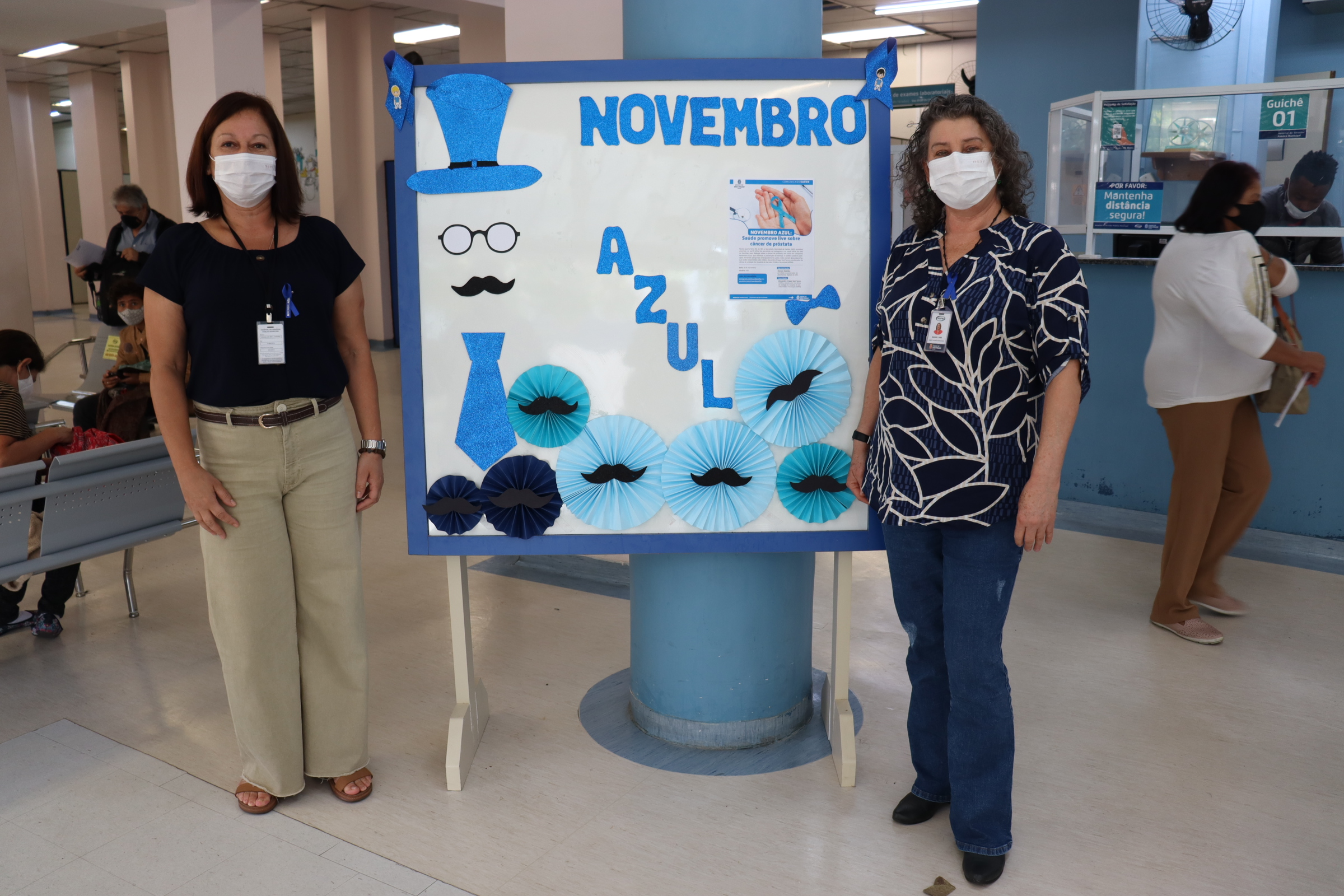 Foto com duas funcionárias, em pé, ao lado do banner escrito Novembro Azul, no saguão principal do hospital